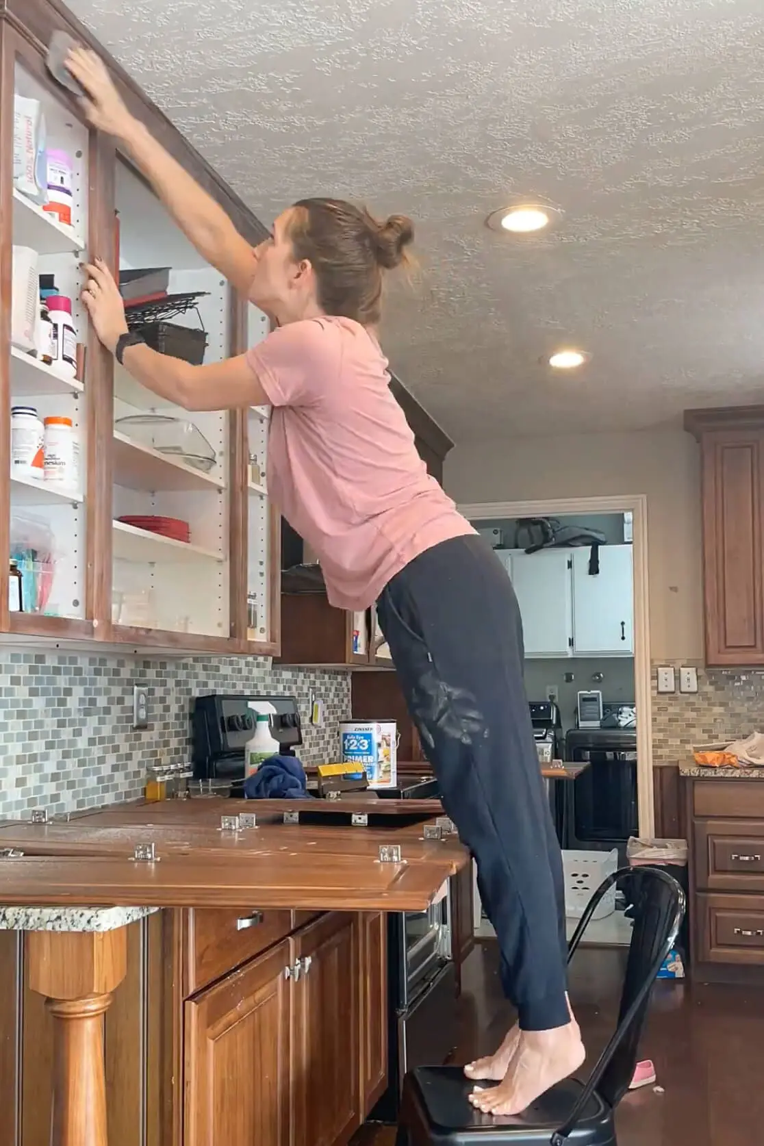 A woman standing barefoot on a chair while sanding the top of a kitchen cabinet. The cabinet doors are removed, and the kitchen counter is covered with tools and supplies, showcasing a hands-on DIY kitchen renovation project.